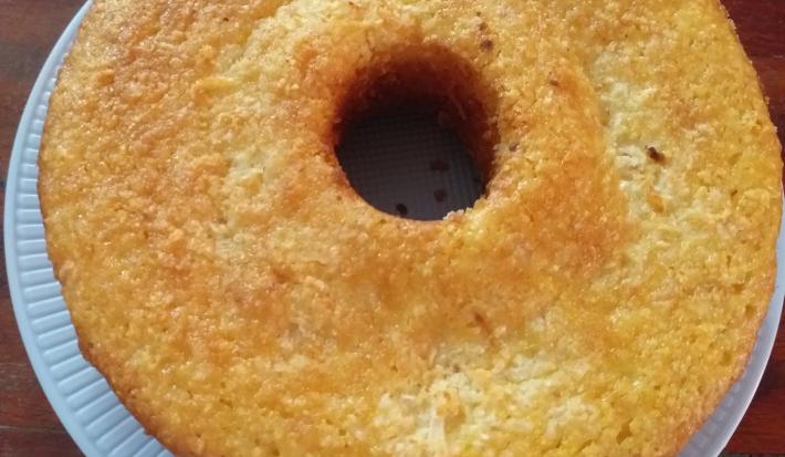 Fotografia em tons de bege em uma bancada de madeira escura, um prato branco redondo e o bolo de milho por cima..