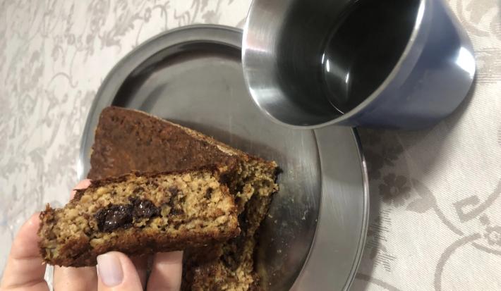 Fotografia de uma mão feminina segurando um pedaço de bolo de banana com aveia e chocolate. A outra parte do bolo está atrás da mão, sobre um recipiente oval de alumínio, ao lado de um copo de aluminio com café.