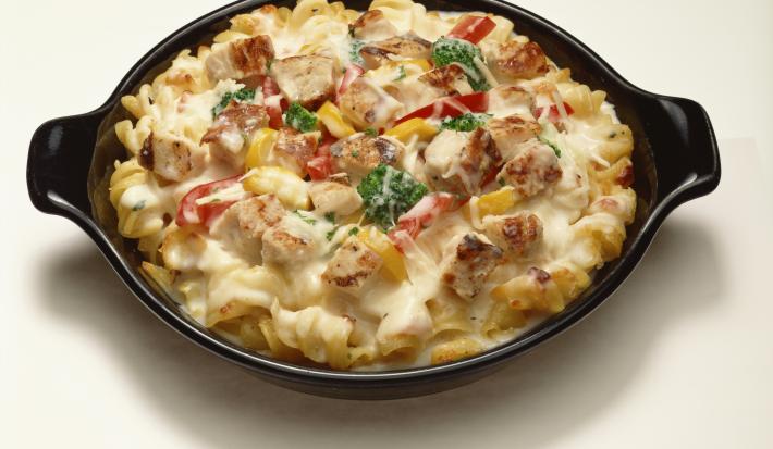 Fotografia de um macarrão cremoso, tipo parafuso, com creme de leite, tomate e manjericão. A pasta está dentro de um recipiente de vidro na cor preta, que está sobre uma mesa branca.