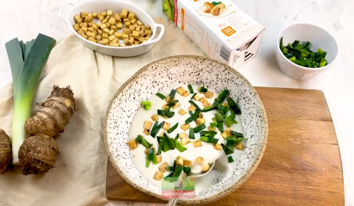 Foto vista de cima de uma mesa com a receita de creme de alho poró com inhame, num prato de cor clara, sobre uma tábua de madeira. Na mesa há potes com croutons, ervas e uma embalagem de bebida vegetal Nature's Heart