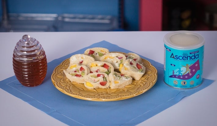 Fotografia em tons claros de um pano azul e quadrado sobre uma mesa de tom claro. Sobre esse paninho tem um prato desenhado amarelo, de vidro, com os sushis de frutas. Ao lado do prato tem a lata de ascenda e um vidro com mel.