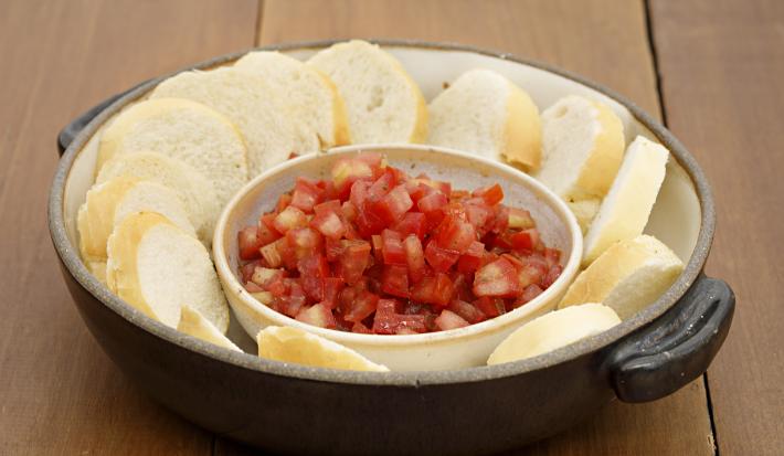 Fotografia em tons de vermelho em uma bancada de madeira com uma panela preta de ferro com alças laterais e vários pães fatiados com um potinho de molhinho de tomate.