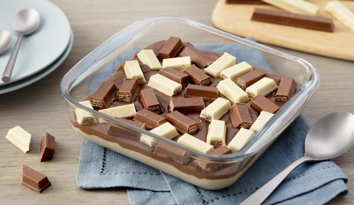 Fotografia em tons de azul em uma bancada de madeira com um pano azul, um refratário de vidro quadrado com o doce de chocolate branco e ao leite dentro, com KitKat ao leite e branco para decorar.