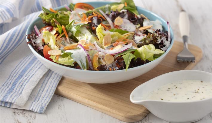 Fotografia em tons de branco e azul de uma bancada branca e uma tábua de madeira, sobre ela um recipiente redondo com a salada e um garfo. Ao lado uma molheira branca com o molho e um paninho branco e azul.