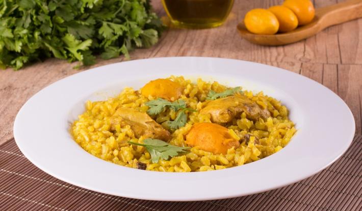 Fotografia em tons de amarelo, em uma bancada de madeira de cor marrom. Ao centro, um prato branco redondo contendo o arroz com frango e pequi. Ao fundo, uma colher de pau contendo alguns pequis e ao lado alguns temperos verdes espalhados.