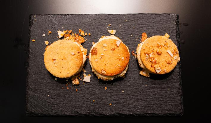 Fotografia em tons de marrom com um prato escuro ao centro. Dentro do prato existe 3 biscoitos recheados com Sorvete de abacaxi com coco