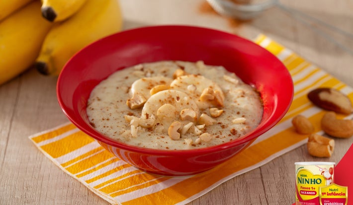 Mingau cremoso servido em tigela vermelha, com banana e castanhas picadas por cima, sobre guardanapo amarelo, em mesa clara.