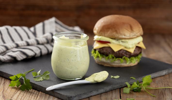 Fotografia em tons de verde em uma bancada de madeira clara, um pano branco com listras pretas, uma tábua de ferro preta e em cima dela, um potinho de vidro com a maionese verde caseira e ao lado um hambúrguer para acompanhar essa deliciosa maionese.