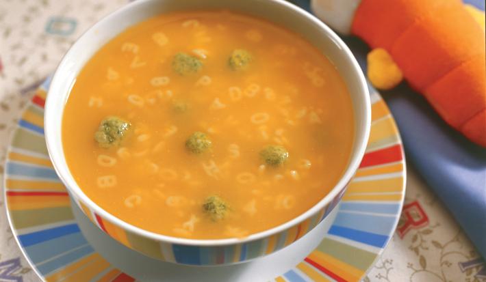 Fotografia em tons de laranja em uma mesa com uma toalha de folhas, um prato raso colorido, um prato fundo redondo colorido com a sopa divertida com bolinhas de espinafre.