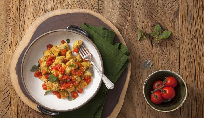 fotografia em tons de marrom e verde de uma bancada marrom vista de cima. Contém um recipiente branco e redondo com Nhoque de Abóbora e molho de tomate com um garfo ao lado para servir. Abaixo um pano verde e ao lado um pote redondo com tomates.