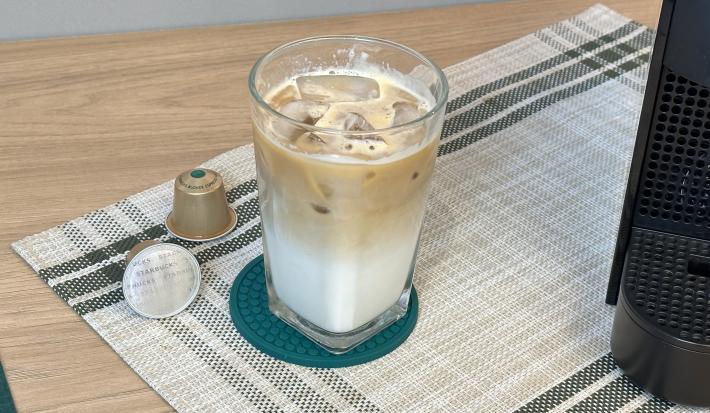 Fotografia em tons de verde escuro em uma bancada de madeira clara, um paninho verde e cápsulas de starbucks ao lado.