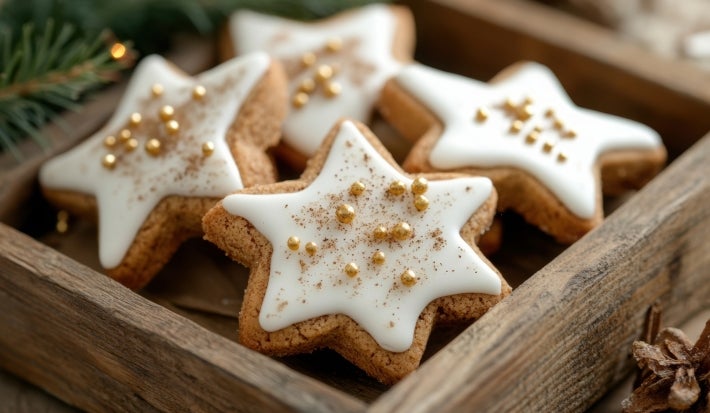 Em cima de uma mesa, biscoitos de estrelas, decorados com glacê branco e confeitos dourados de bolinha.