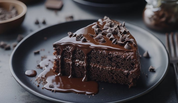 Fatia de bolo de chocolate em camadas, recheada e coberta com ganache bem cremosa. Por cima, gotas de chocolate dão um toque especial. A cobertura escorre pelo prato, deixando a apresentação ainda mais apetitosa.