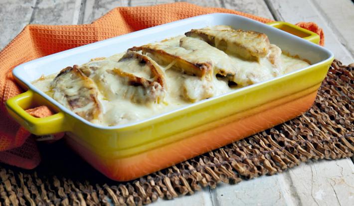 Fotografia em tons de laranja, amarelo e branco de uma bancada de madeira branca com um apoio de mesa de madeira e um paninho laranja, sobre eles um recipiente retangular laranja e amarelo com o peixe á delicia.
