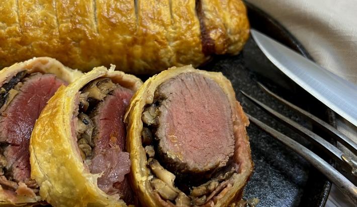 Foto em tons de amarelo da receita de filé wellington servida em três cortes e uma peça inteira e sobre uma tábua de pedra preta com talheres prateados ao lado direito