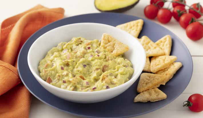 Fotografia em tons de azul em uma bancada de madeira clara com um pano laranja ao lado e um prato grande azul e um prato fundo branco com o guacamole dentro. Ao lado, um pedaço de avocado, alguns tomatinhos e biscoitinhos.