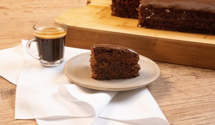 Fotografia de uma bancada de madeira, e sobre ela tem uma torta de chocolate com café em um prato de vidro em tom pastel, que está apoiado sobre um pano branco. Ao lado da sobremesa tem uma xícara com Nescafé, também sobre o pano.