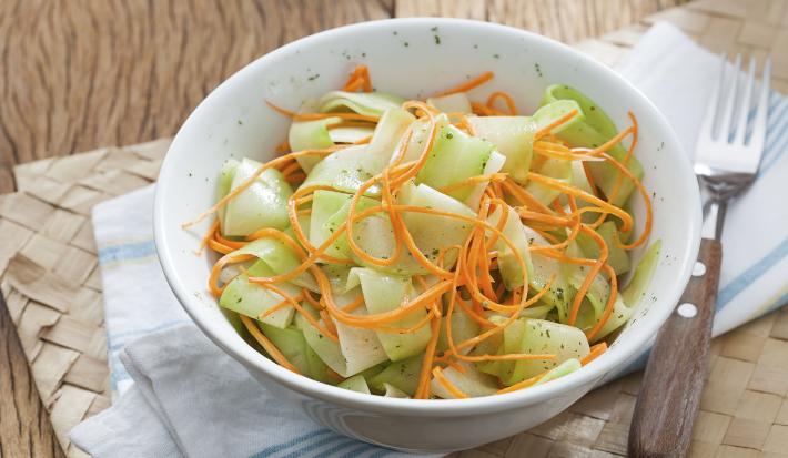 Fotografia em tons de verde em uma mesa de madeira com um pano branco com detalhes em azul e um prato fundo branco com a salada de chuchu dentro.