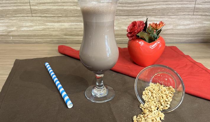 Fotografia em tons de vermelho e marrom com uma taça ao centro. Dentro da taça existe uma bebida cremosa de leite com NESCAU Extra Cacau e Iogurte. Ao lado existe um pote com amendoins