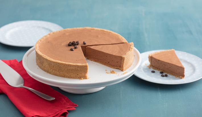 fotografia em tons azul e branco. Contém três pratos brancos, um deles esta com uma fatia de torta e o outro com a torta para servir. Ao lado um pano vermelho com uma espátula por cima. O terceiro prato esta vazio.
