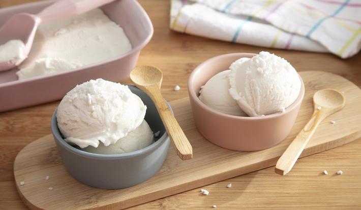 Foto em tons claros da receita de sorvete de coco cremoso caseiro servida em duas porções em potinhos azul e rosa com colheres de madeira sobre uma tábua de madeira com a receita maior ao fundo