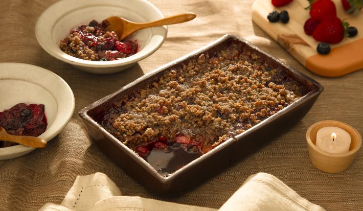 Foto em tons de marrom da receita de doce de aveia com frutas vermelhas servida em uma forma de madeira marrom escura com dois pratos fundos brancos ao lado com porções do doce