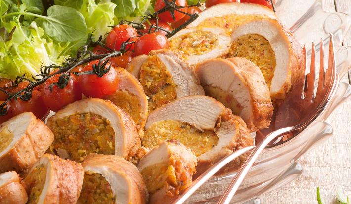 Fotografia em tons de verde e bege em uma mesa de madeira clara com um prato fundo de vidro com os filés de frango cortados ao meio recheados de farofa e salada de alface e tomate servindo como acompanhamento.