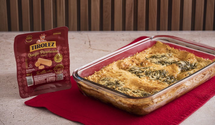 Foto da receita de Lasanha e Queijo Parmesão e Pesto, servida em uma travessa de vidro, sobre um pano vermelho. Ao lado dela há uma embalagem de Queijo Parmesão Tirolez