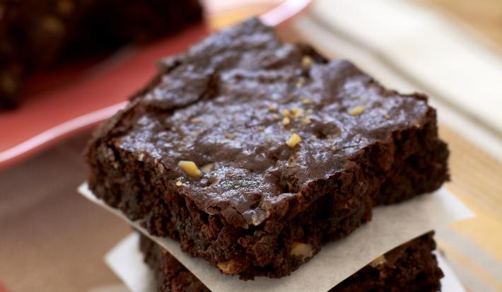 Fotografia em tons de marrom e branco de uma bancada de madeira clara e um paninho marrom, sobre ela fatias de brownie separadas por papel manteiga. Ao fundo um prato vermelho com pedaços de brownie.