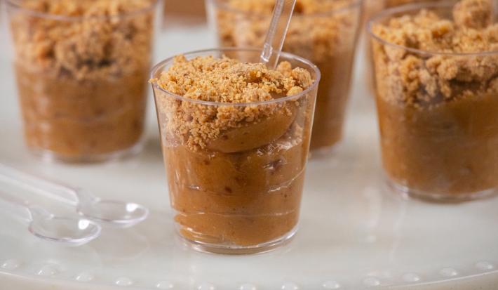 Fotografia em tons de branco com vários potinhos de plástico ao centro. Dentro de cada potinho existe um brigadeiro de cor caramelo feito com paçoca e coberto com paçoca triturada.