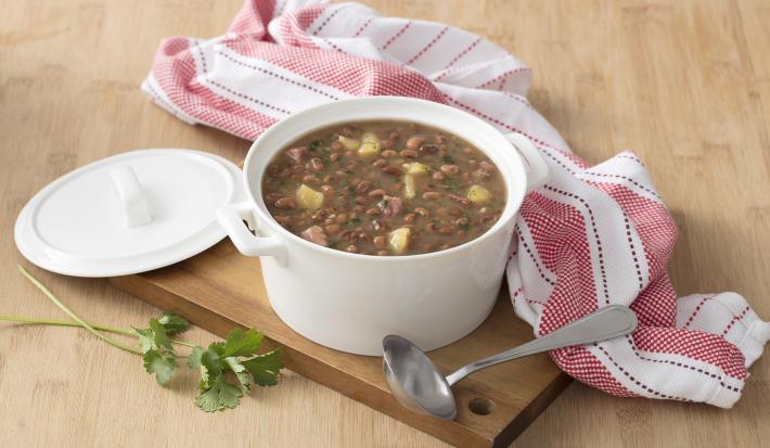 Fotografia em tons de marrom, branco e rosa de uma bancada de madeira marrom vista de cima, contém uma tábua de madeira com uma panela redonda branca com alças e feijão dentro com pedaços de carne-seca e mandioca. Ao lado a tampa da panela e um pano.