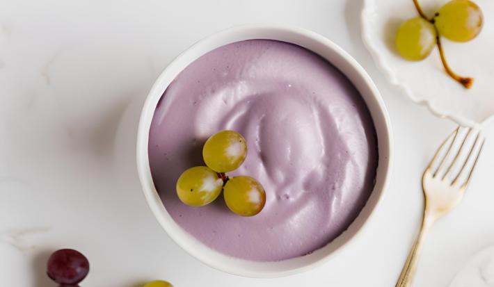 Fotografia em tons de branco e roxo com um pote branco ao centro. Dentro do pote existe uma receita de mousse de uva de cor roxa com algumas uvas verdes de enfeite.
