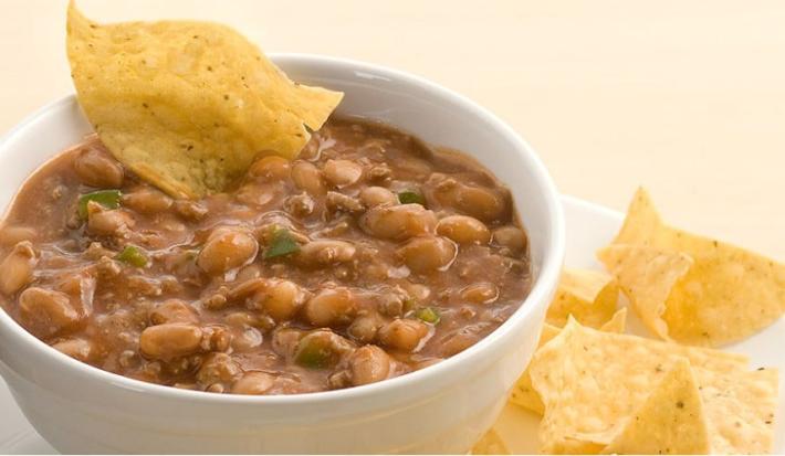 Fotografia de um bowl branco com chilli dentro, grãos de feijão da superfície e servido com tortilhas de milho ao lado no prato branco.