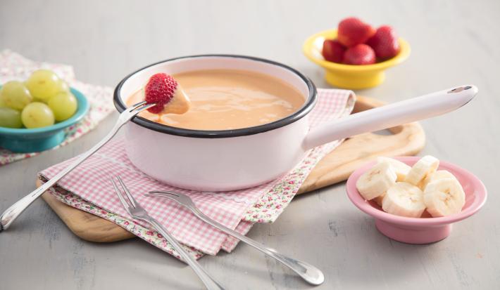 Fotografia em tons de branco, caramelo e rosa, tendo ao centro panela em ágata contendo doce de leite cremoso, garfinhos para fondue ao lado sobre guardanapo quadriculado e peça de madeira, no entorno potinhos com frutas picadas, tudo sobre bancada cinza.