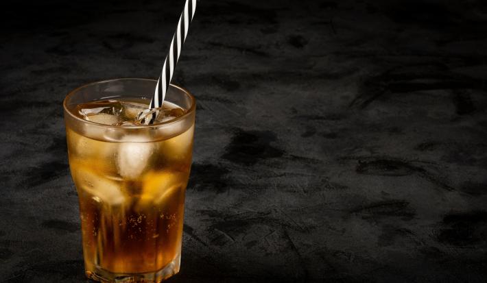 Fotografia em tons escuros de um copo de vidro com uma bebida de guaraná com cubos de gelo e um canudo preto e branco dentro do copo, o qual está sobre uma mesa preta.