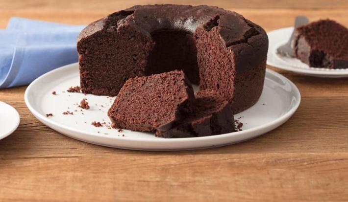 Fotografia em tons de marrom e branco de uma bancada de madeira clara com um paninho azul claro e um prato branco redondo com o bolo de chocolate. Aos lados dois pratos brancos pequenos com fatias de bolo de chocolate e um garfo.