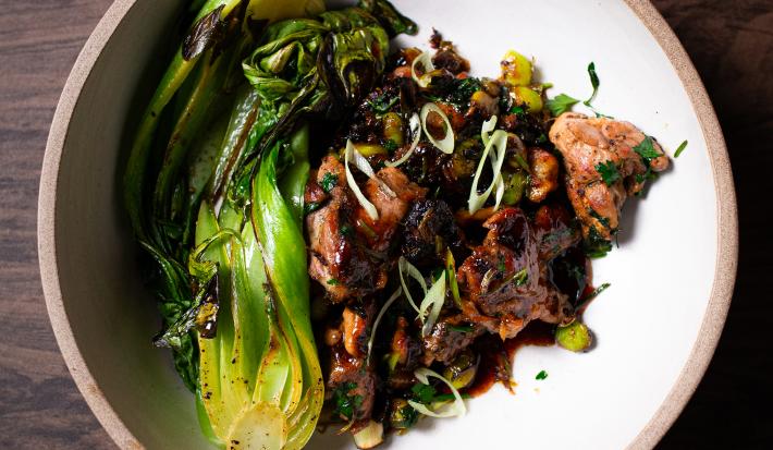 Fotografia em tons de marrom e cinza com um prato cinza ao centro. Dentro do prato existe uma receita de ragu de pato acompanhado de cogumelos, edamames e bok choy