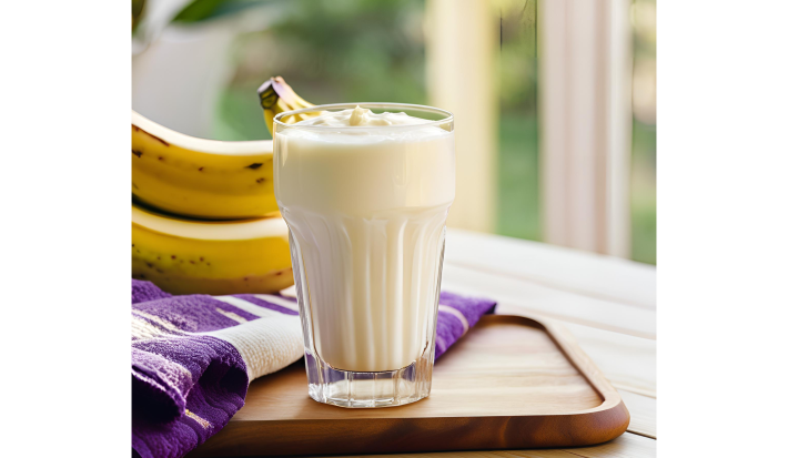 Fotografia em tons de roxo e amarelo com um copo de vidro ao centro. Dentro do copo existe um shake de coco, batata doce e banana de cor branca. Ao fundo existe duas bananas