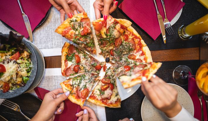 Fotografia vista de cima de uma pizza com escarola, linguiça, muçarela de búfala e manjericão com tomates. A pizza está sobre um prato raso branco, cortada em 8 fatias. Na imagem, há quatro pessoas estão puxando uma fatia, e ao redor, copos e talheres.