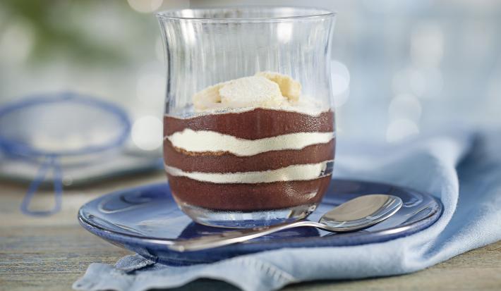 Fotografia em tons de azul e cinza, em uma bancada contém um pano azul e em cima um prato com um copo que contém açaí, banana e leite em pó. Ao lado uma colher para servir.