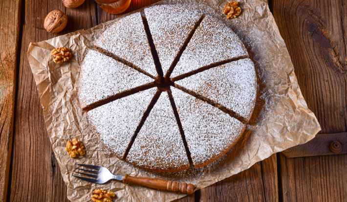 Foto em tons de marrom da receita de bolo alemão de mel servida cortada em 8 pedaços polvilhada com açúcar de confeiteiro, bolo em cima de uma mesa de madeira com nozes ao lado