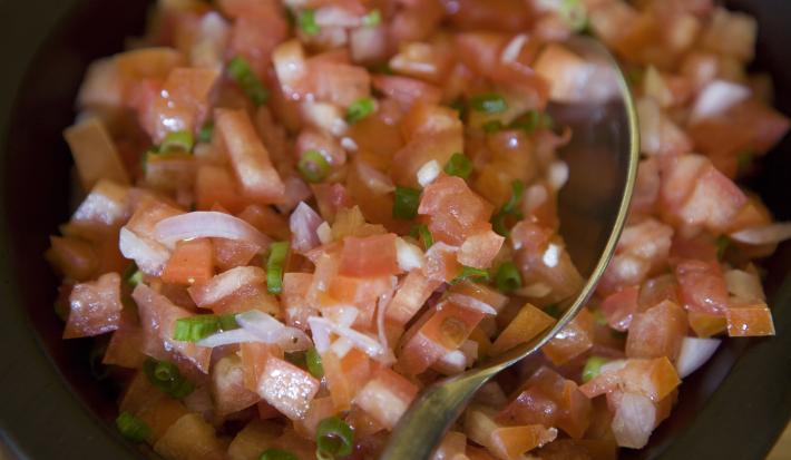 Fotografia de um molho vinagrete com cebola e salsinha, e uma colher apoiada na salada. A vinagrete está em um recipiente pequeno e preto.