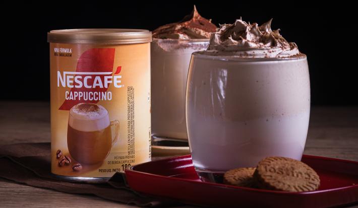 Foto da receita de cappuccino super cremoso servida em dois copos de vidro sobre uma louça vermelha em cima de uma mesa de madeira