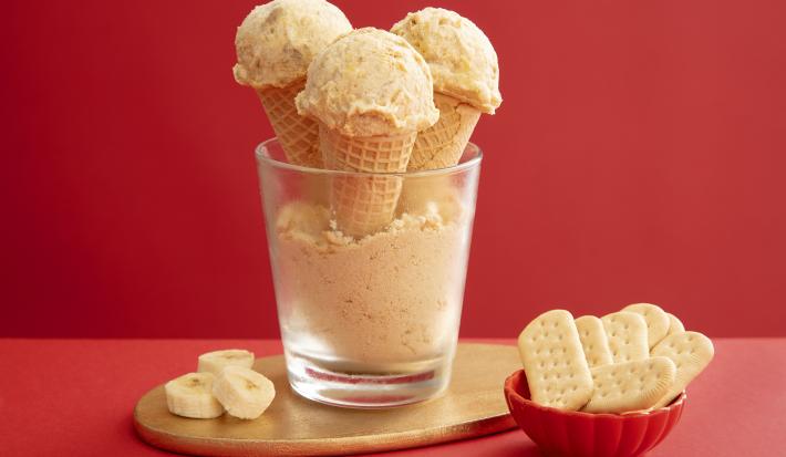 Foto da receita de Sorvete de Banoffee. Observa-se um copo de vidro com farofinha de biscoito no fundo e três casquinhas de sorvete com uma bola dispostas no copo. Ao lado direito, um potinho com biscoitos e, ao esquerdo, rodelas de banana