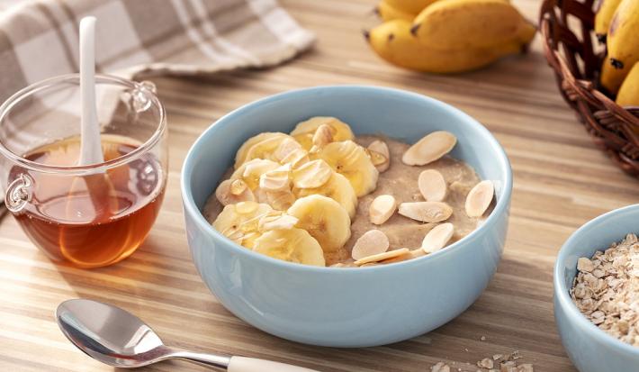 Fotografia em tons de branco em uma bancada de madeira de cor marrom. Ao centro, há uma tigela azul com o mingau, ao lado há um pote com mel e ao fundo, um pano cinza quadriculado, bananas e aveia.