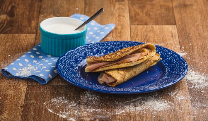 Imagem em tons de marrom e azul claro e escuro. No centro há um prato azul com duas quesadillas recheadas e, ao lado esquerdo, há um tecido azul com bolinhas brancas e, sobre ele, um pote azul com o creme azedo e uma colher.
