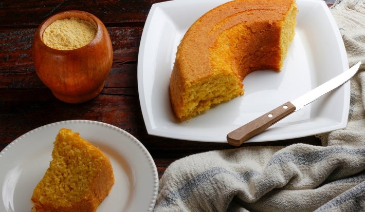 Fotografia de um bolo de fubá parcialmente fatiado em um prato branco, com uma crosta dourada e interior amarelo. Um recipiente de madeira com fubá está ao lado.