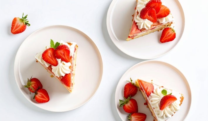 Fotografia em tons claros de três pratos brancos sobre uma mesa off white. Dentro dos pratos rasos tem uma fatia da sobremesa, super consistente, com pedaços de morango em cada prato.