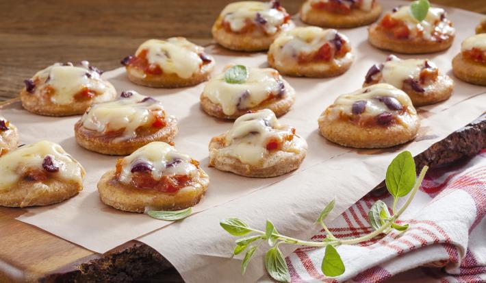 Fotografia em tons de marrom em uma bancada de madeira com uma tábua de madeira escura ao centro com muitas mini pizzas de frigideira apoiadas nela. Embaixo, um pano branco com listras vermelhas.