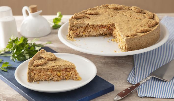 Receita da Torta de Frango sem Glúten. Observa-se a torta em um prato grande branco e, ao lado esquerdo, um prato individual com uma fatia. Como decoração da foto, tem-se um guardanapo listrado azul e uma espátula de cortar.